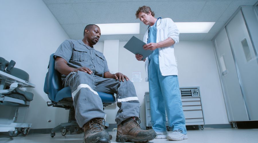 Steps to Take After a Workplace Injury in Orange County 2 Workplace injury victim seated in an urgent care exam room, doctor reviewing chart, patient wearing work clothes, clean medical environment.