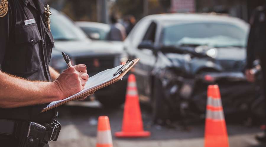 A Costa Mesa police officer documenting a car accident scene, taking notes beside damaged vehicles parked roadside. Traffic cones and clipboard visible.
