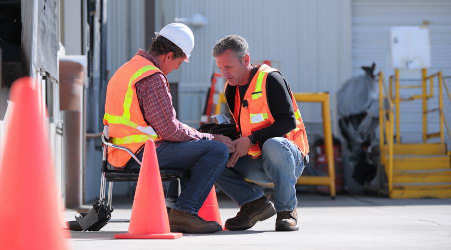 Steps to Take After a Workplace Injury in Orange County 1 Workplace injury scene in Orange County, employee seated on a loading dock receiving first aid from a coworker, warehouse environment, safety cones nearby