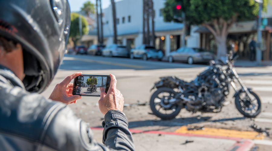 Motorcycle Accidents in Southern California: Legal Rights Explained 2 a motorcyclist using a smartphone to photograph vehicle damage and road conditions after a minor crash on a California street, motorcycle parked safely to the side, natural daylight