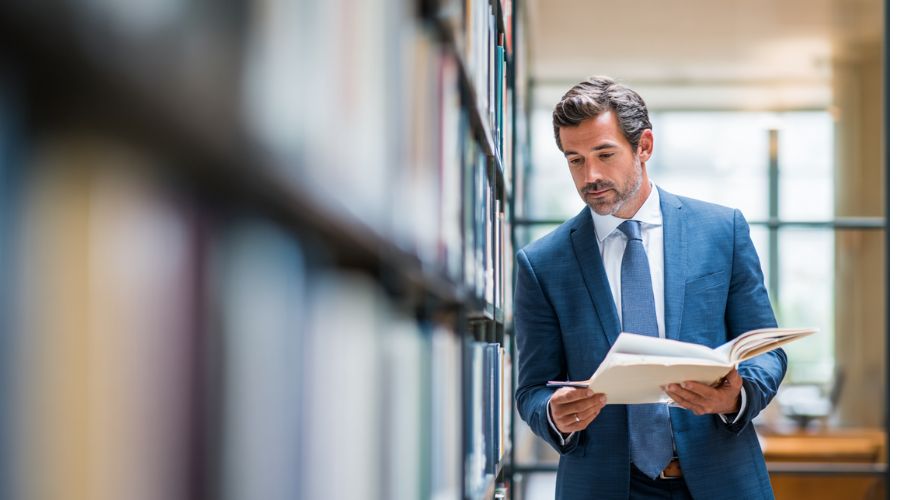 Un abogado profesional de pie en una oficina de abogados moderna, sosteniendo un expediente abierto y revisando documentos con una expresión concentrada y pensativa, estanterías de piso a techo borrosas en el fondo, una suave luz natural de ventana y vestimenta de negocios neutral.