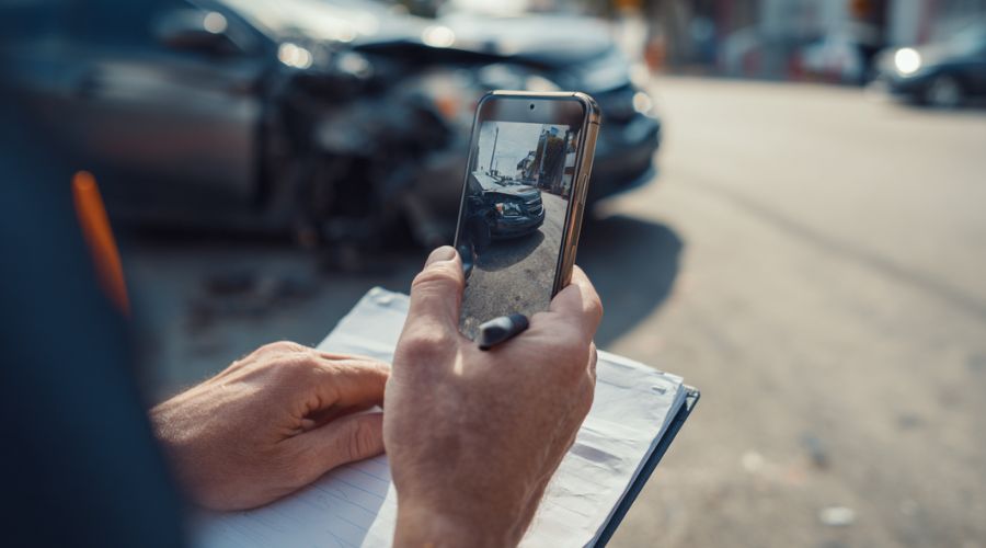 Qué hacer después de un accidente automovilístico en Anaheim, CA 2 Una persona junto a un coche averiado en Anaheim, usando un teléfono inteligente para fotografiar los daños. Bloc de notas visible en la mano, postura tranquila.