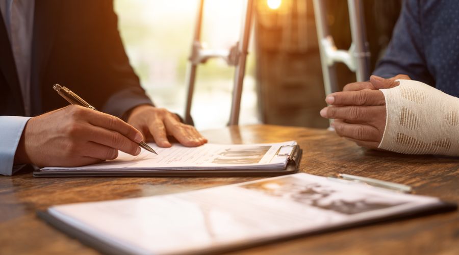 A realistic image of a personal injury lawyer reviewing accident photos and medical records with a client at a law office table. The client wears a brace and uses crutches. Warm lighting, professional atmosphere.