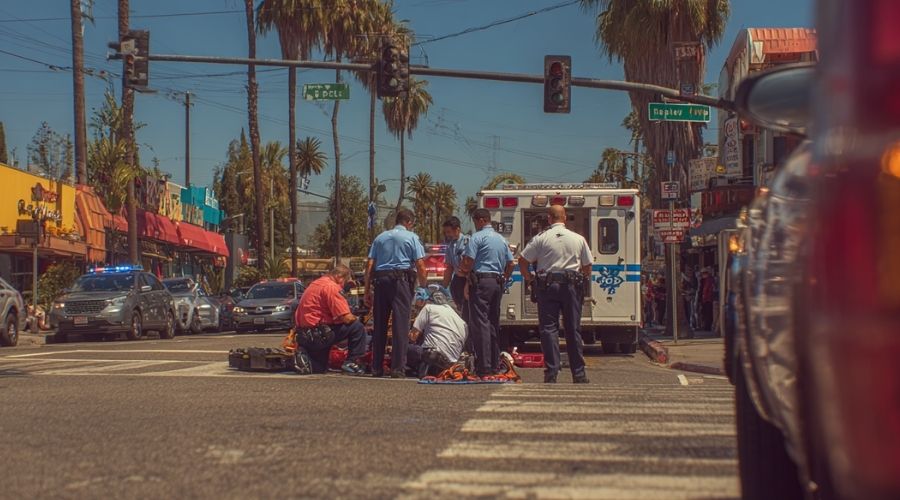 Fotografía ultrarrealista de un accidente de peatones en una concurrida intersección de Santa Ana: una persona en el suelo siendo asistida por los servicios de emergencia, un coche detenido cerca, policía y ambulancia en el lugar, paso de peatones y semáforos visibles, entorno urbano del sur de California con palmeras, luz natural brillante, estilo cinematográfico documental.