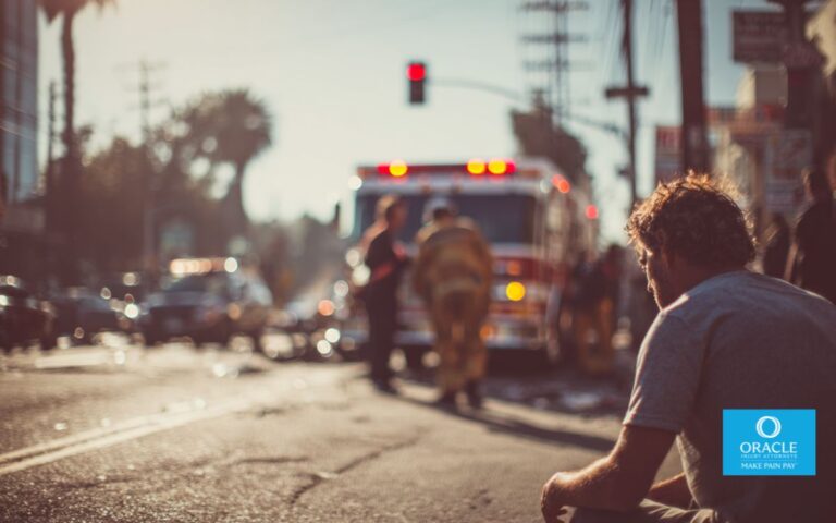 A serious car accident scene in an urban Southern California setting, injured person sitting on the curb holding their arm while paramedics assist nearby, damaged vehicles in background, natural daylight, realistic photography style, emotional but not graphic, no text