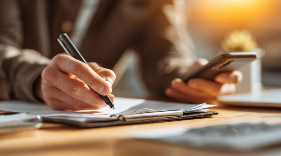 Murrieta Work Accidents: What Happens If Your Employer Doesn’t Report Your Injury? 2 Close-up of a person organizing documents, taking notes, and saving messages on a smartphone, desk with medical records and notepad, bright focused lighting, sense of action and control, realistic modern workspace