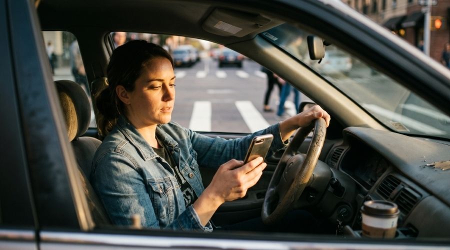 Un conductor mirando su teléfono inteligente mientras está al volante en un paso de peatones, poca profundidad de campo, iluminación lateral dramática, estilo fotográfico editorial, sin texto.