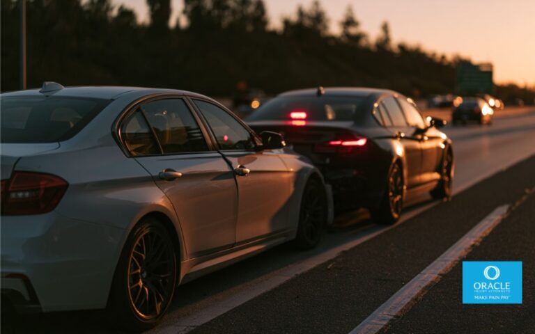 A cinematic wide-angle photograph of a rear-end car collision on a California freeway at golden hour. Two modern sedans, front car stopped, rear car slightly crumpled. Emergency hazard lights blinking. Blurred traffic in the background. Shot from a low angle, shallow depth of field, moody warm tones with long shadows. No visible injuries, no gore. Editorial photography style, Canon EOS R5, 35mm lens, f/2.8.