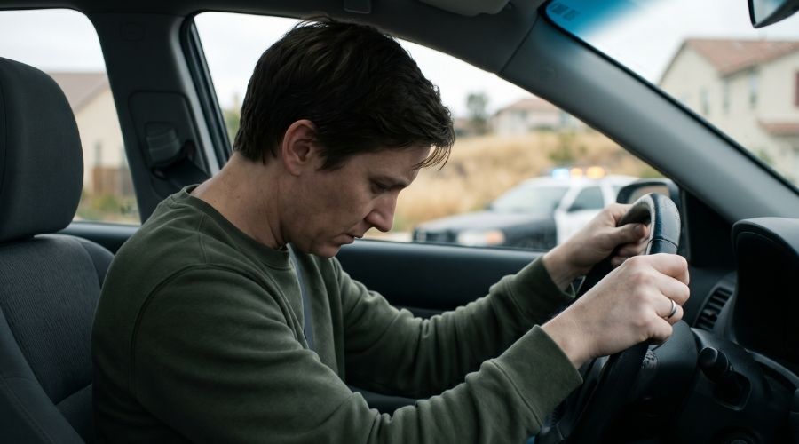 Close-up photorealistic image of a person sitting in a car driver's seat, hands gripping the steering wheel tightly, head slightly bowed. Visible tension in the posture but no external injuries. Soft natural light coming through the windshield. Blurred background of a suburban California street. Conveys shock and adrenaline aftermath. Cinematic, muted cool tones. Shot with a 50mm lens, shallow depth of field.