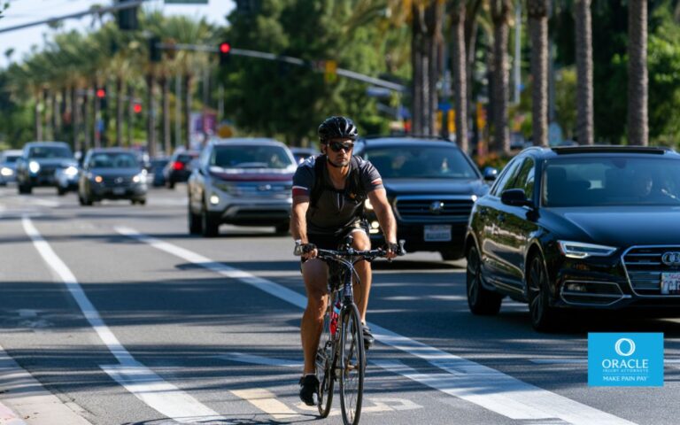 A highly realistic daytime scene of a cyclist riding confidently in a designated bike lane on a busy Orange County boulevard. Vehicles pass while maintaining a safe three-foot distance, with visible road markings, palm trees, traffic signals, and clear California weather. The cyclist wears a helmet and reflective clothing. Professional editorial photography, ultra-detailed, natural lighting, 16:9, no text, no logos.