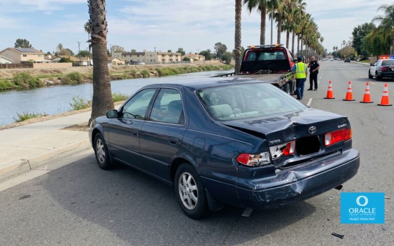 A car with visible rear-end collision damage on a suburban Riverside street, daylight, police cones in the background, no graphic injuries shown.