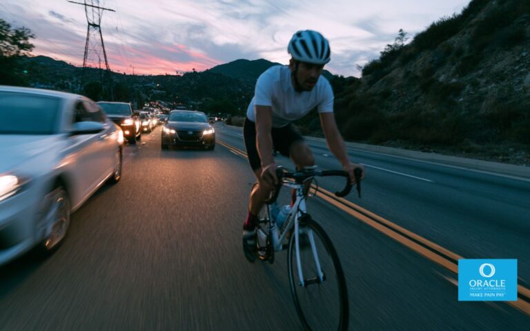 A cyclist riding along a Riverside County road at dusk with cars passing nearby, realistic photography style, wide-angle shot emphasizing the vulnerability of cyclists on busy streets.