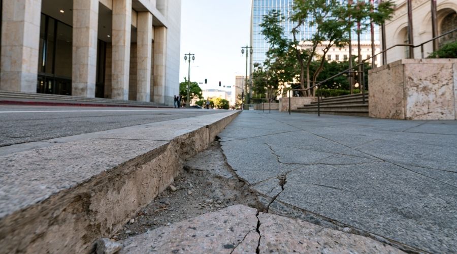 Slip and Fall Claims: Proving Property Owner Negligence in California 4 A wide shot of a public sidewalk with a visibly cracked or uneven pavement section near a government building or city street, daylight, no people mid-fall. Represents the government entity liability and six-month deadline angle.