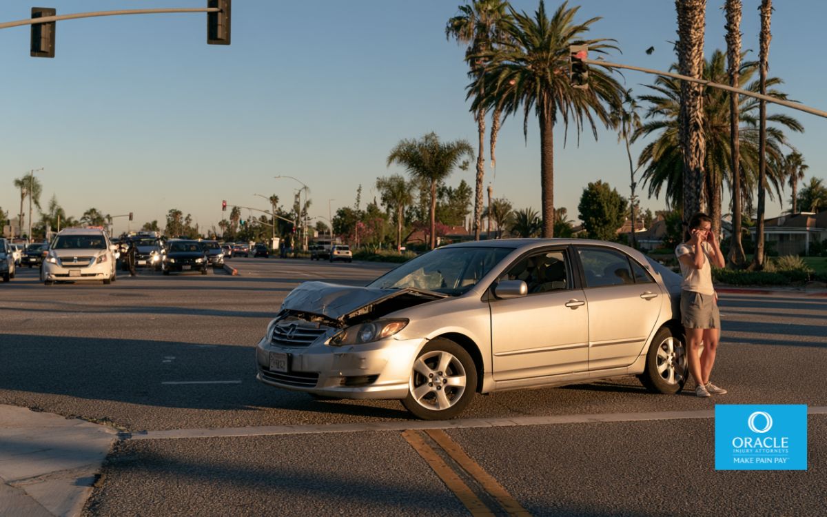 Fotografía muy realista de un sedán plateado dañado detenido en medio de una intersección de California momentos después de un accidente con fuga. El tráfico continúa en segundo plano mientras que el vehículo culpable ya no es visible. El conductor está de pie junto al auto haciendo una llamada a los servicios de emergencia. Iluminación de la hora dorada, palmeras, suburbio del sur de California, composición cinematográfica, fotografía de estilo documental, ultra detallada, colores naturales, sin texto, sin logotipos.,