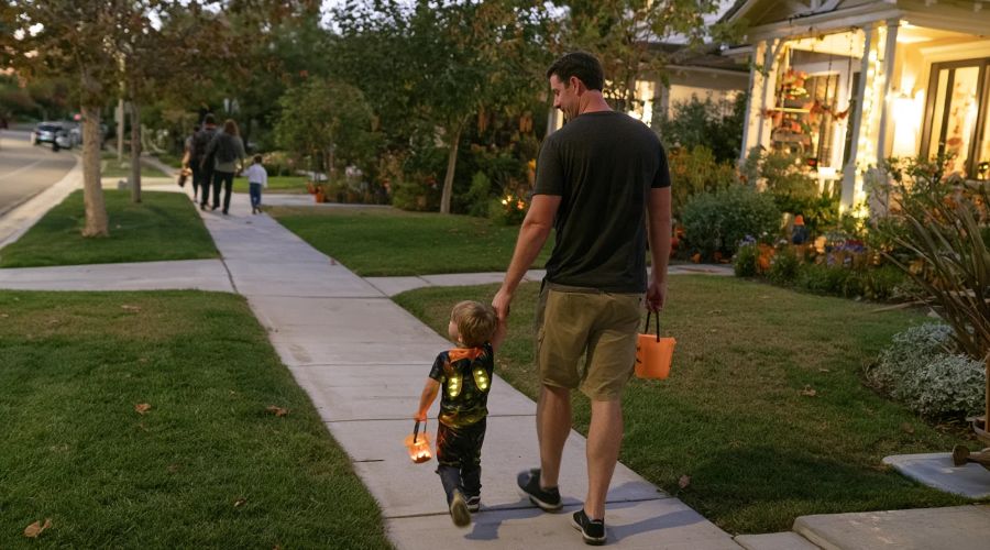 Parent holding a child's hand while trick-or-treating on a well-lit sidewalk in a California neighborhood.